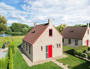 Ein zweistöckiges Gebäude mit roter Tür und Fenstern, umgeben von einer gepflegten Rasenfläche und einem Gewässer.