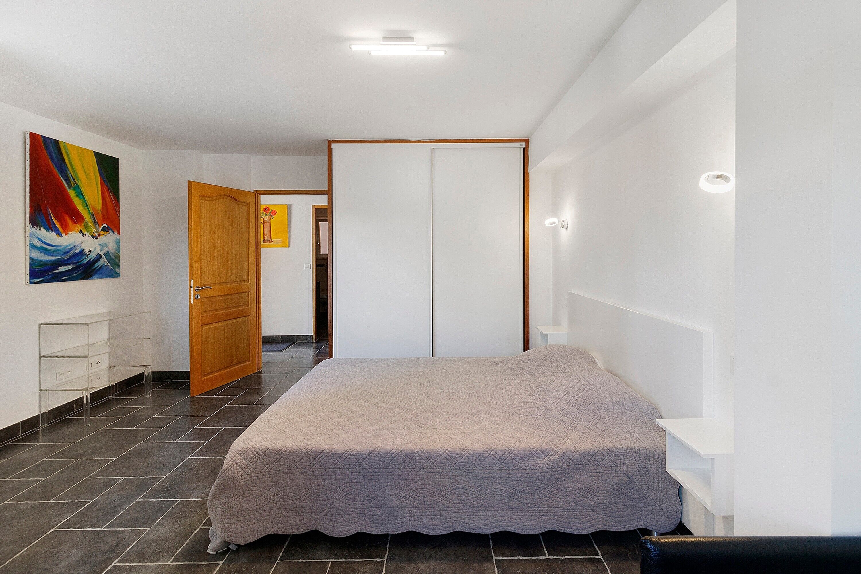 A bedroom with a bed, a door, a wall-mounted shelf, and a painting.