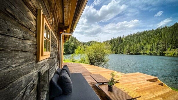 A wooden cabin with a deck, a sofa, and a view of a lake and forest.