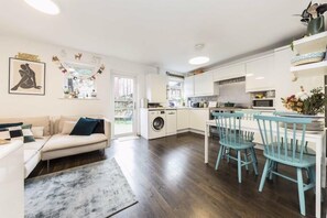 A modern kitchen with white cabinets, a dining area with blue chairs, a washing machine, and a sofa with a blue pillow. - Four-bedroom home in Chiswick, West London, near transport links (London)