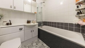 A modern bathroom with a white sink, a toilet, a bathtub, and a shower. There are shelves with toiletries and a decorative plant. - Four-bedroom home in Chiswick, West London, near transport links (London)