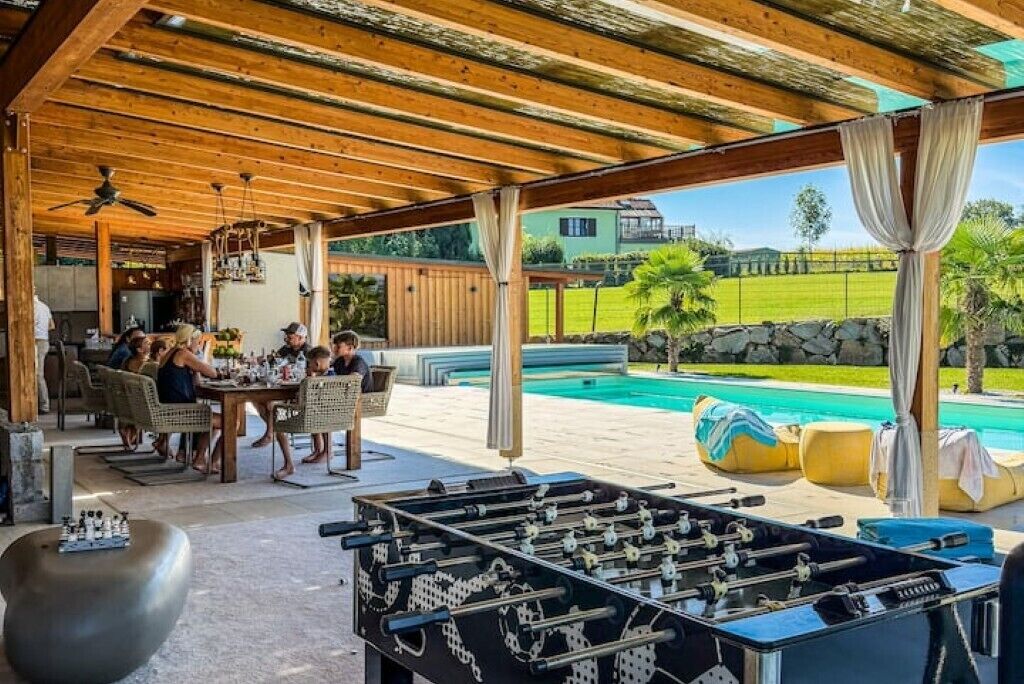 Un espacio exterior techado con mesa de fútbol de mesa, asientos y una piscina al fondo.