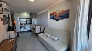 En moderne stue med en hvid sofa, et spiseområde med stole, et køkken med hvide skabe og et stort vindue.