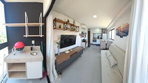 Una sala de estar moderna con sofá, televisión, zona de cocina y un balcón con un objeto rojo.