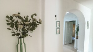 En gang med en grøn vase med blomster på en hvid reol, en dør og en lille potteplante på et hvidt piedestal.