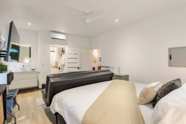 A modern hotel room with a bed, bedside table, and a desk with a chair. There is a wardrobe, an air conditioning unit, and a ceiling fan.