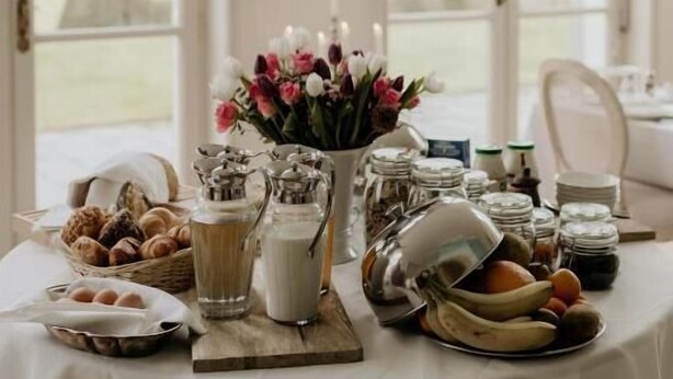 Mesa posta com comida e bebidas, um vaso de flores, e vista para o exterior através de grandes janelas.