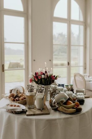 A table set with food and drinks, a vase of flowers, and a view of the outdoors through large windows. - B&B Kasteel Den Tip (Arendonk)