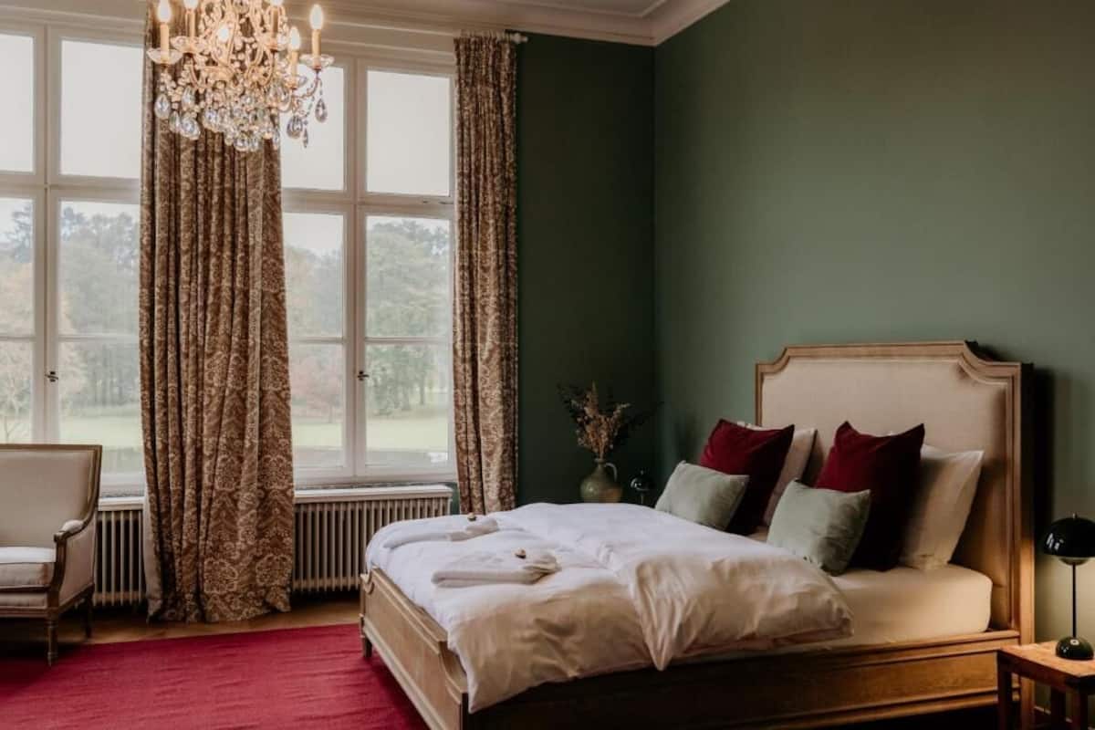 A bedroom with a large bed, a chair, a chandelier, and a view of the outdoors through a window.