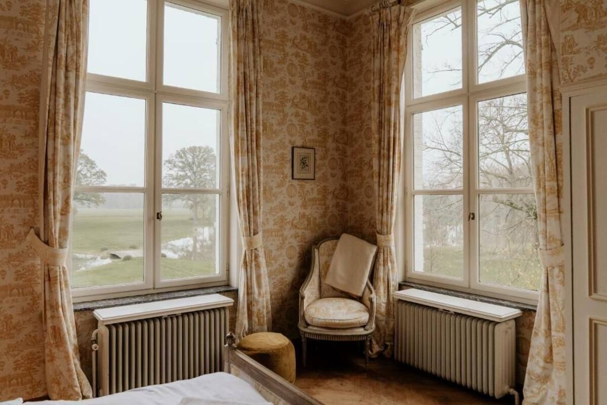 A room with two large windows, a radiator, a chair, and a bed.