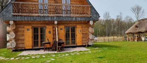 A log cabin with a balcony, a stone patio, and a thatched-roof structure in the background.