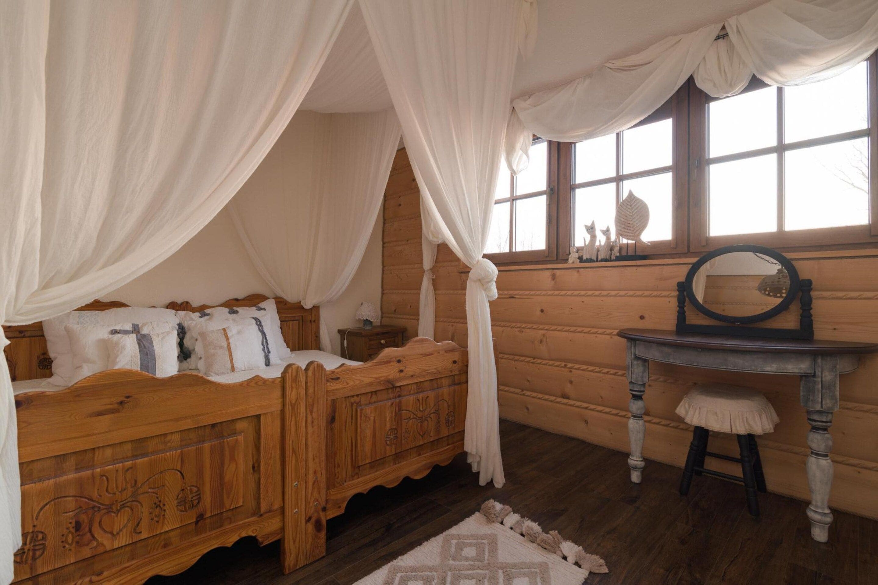 Ein hölzernes Bett mit Baldachin, ein Nachttisch, eine Kommode, ein Spiegel und ein Fenster mit Blick ins Freie.
