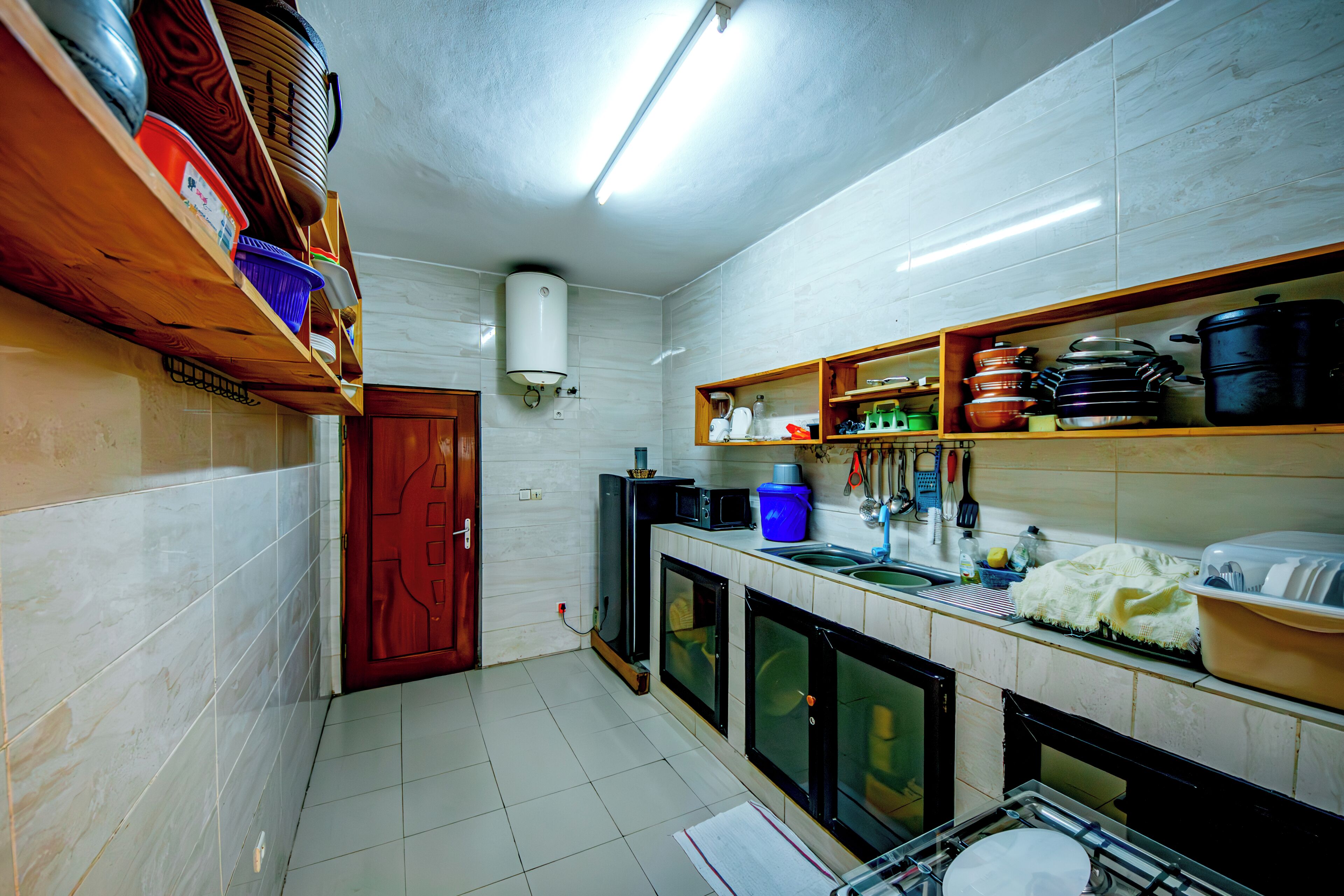 A compact kitchen with white tiled walls, a wooden door, and shelves holding kitchenware.