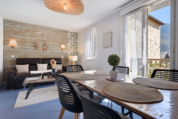A modern living room with a dining table, black chairs, a sofa, and a large window with a view of mountains.