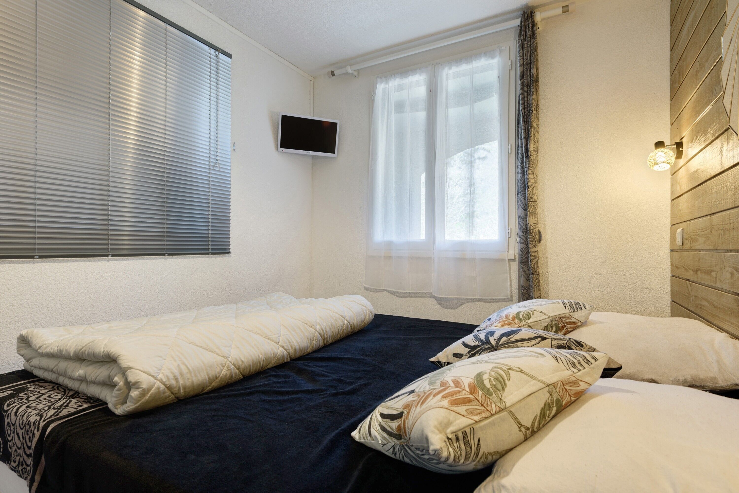 Une chambre à coucher avec un lit, un téléviseur fixé au mur et une fenêtre avec des rideaux.