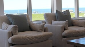 Ein Zimmer mit zwei beigen Sesseln und einem Holztisch, große Fenster mit Blick auf das Meer und Gebäude.