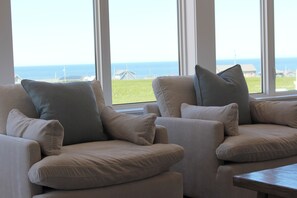Ein Zimmer mit zwei beigen Sesseln und einem Holztisch, große Fenster mit Blick auf das Meer und Gebäude.