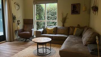 A living room with a brown leather sofa, a round wooden coffee table, a brown armchair, and a large window with a view of trees.