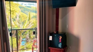 A room with a sliding glass door leading to a view of a valley, a television on a stand, and a red chair. - Pousada Finca dos Anjos (Passa Quatro)