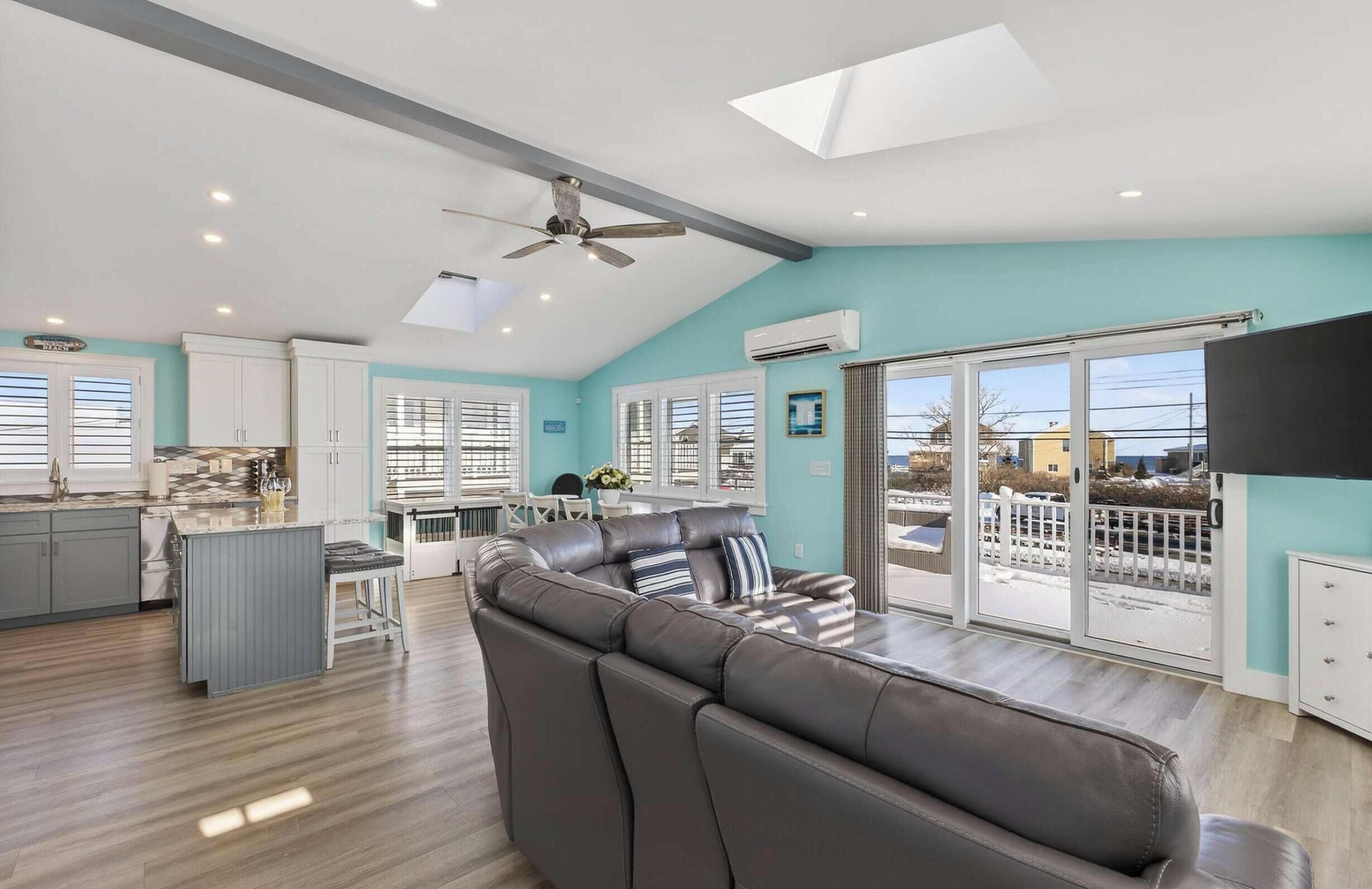 A modern kitchen with white cabinets, a dining area with a table and chairs, a large leather sofa, and a balcony with a view.