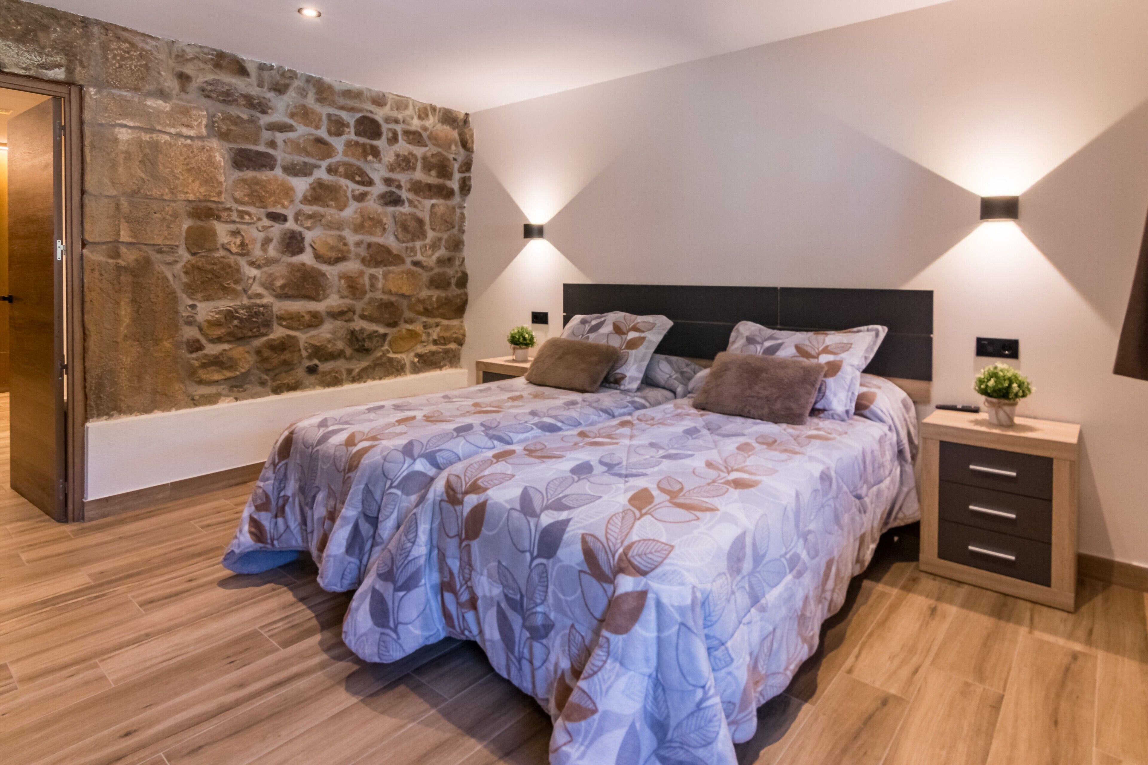 Een slaapkamer met een stenen muur, een bed met een sprei met bladmotief, nachtkastjes met lampen en een houten vloer.