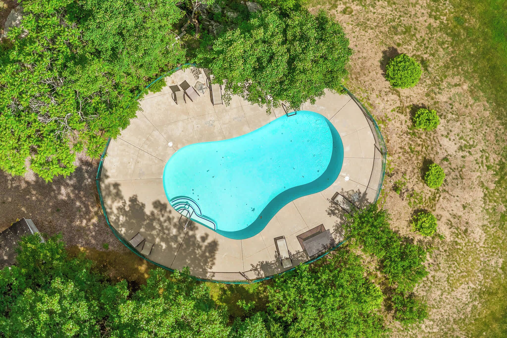 Vue aérienne d’une piscine entourée d’arbres et bordée d’une terrasse en béton avec des chaises longues.