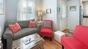 Un espace de vie compact avec un canapé à rayures, un fauteuil rouge, une petite table et un coin cuisine à l’arrière-plan.