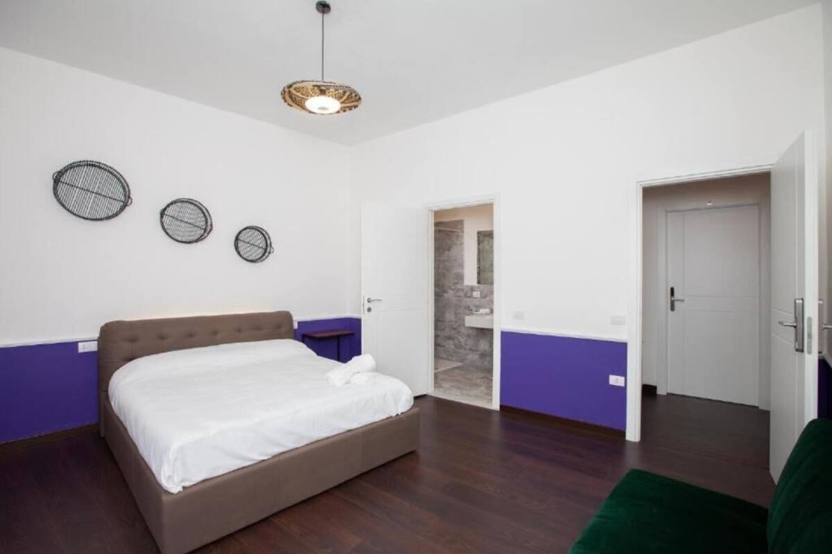 A bedroom with a bed, a nightstand, a wall-mounted light fixture, and a door leading to another room.