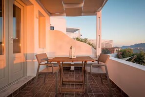A balcony with a table and chairs, a door, and a view of buildings and hills. - Vista d'Oro Luxury Villas (Santorini)