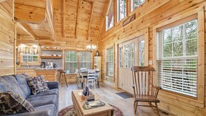 Interieur van een houten chalet met een bank, een eettafel en stoelen, een keuken en grote ramen.