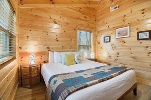 Un dormitorio de cabaña de madera con una cama, almohadas, colcha, mesita de noche, lámpara y cuadros en la pared.