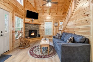 Ein Holzblockhaus-Interieur mit Kamin, einer Couch, einem Schaukelstuhl und einem Deckenventilator.