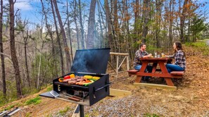 Una parrilla con diversos tipos de carne y verduras, una mesa de picnic y un bosque de fondo.