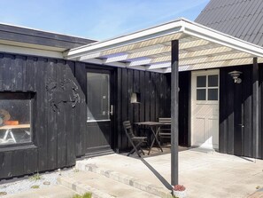 Ein schwarzes Holzgebäude mit überdachter Terrasse, einem Tisch und Stühlen sowie einer Tür mit Glasfenster.