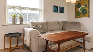 Een moderne woonkamer met een bank, een salontafel, een klein bijzettafeltje en een raam met planten.