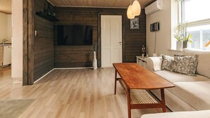 Een moderne woonkamer met een houten plafond, een bank met gekleurde kussens, een houten eettafel en een raam met planten.