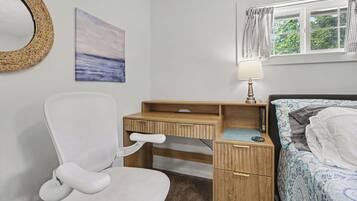 Une chambre avec un bureau en bois, une chaise blanche, un lit recouvert d’une couverture à motifs, une fenêtre avec des rideaux et un miroir fixé au mur.