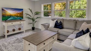 Ein Wohnzimmer mit Fernseher, Sofa, Couchtisch und Fenstern mit Blick auf Bäume.