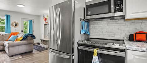 A modern kitchen with stainless steel appliances, a dining area with a sofa, and a view of greenery outside.