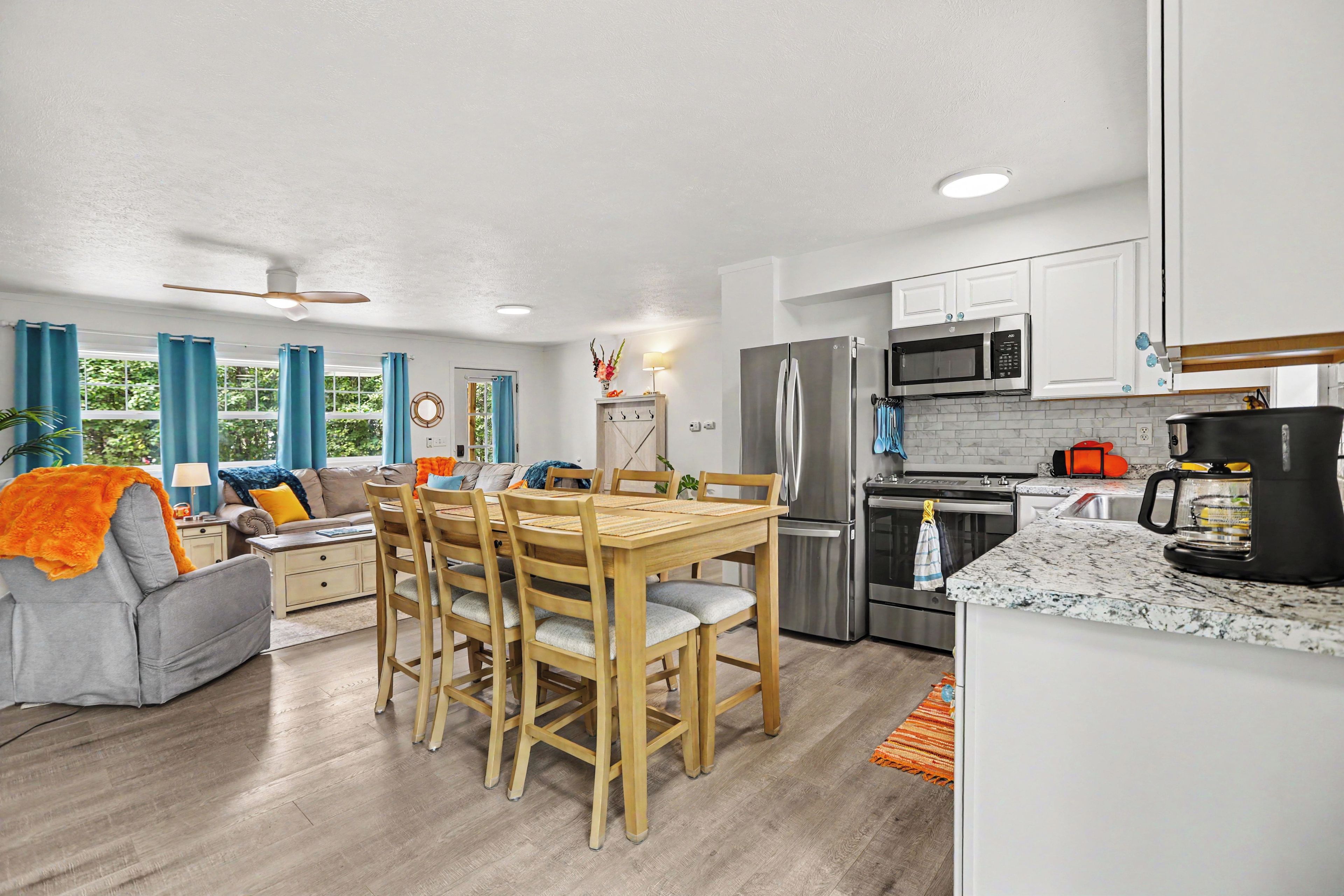A modern kitchen with stainless steel appliances, a dining area with wooden chairs, and a living room with a sofa and curtains.