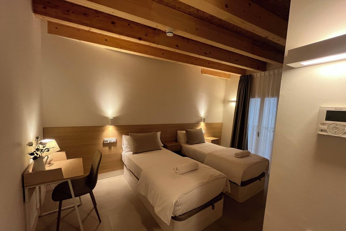 A hotel room with two beds, a desk, a chair, and a thermostat on the wall.