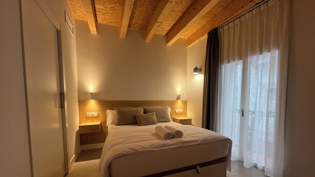 A bedroom with a wooden ceiling, a bed with pillows, a nightstand, and a window with curtains. - HOTEL LUNA NUEVA (Tarragona)