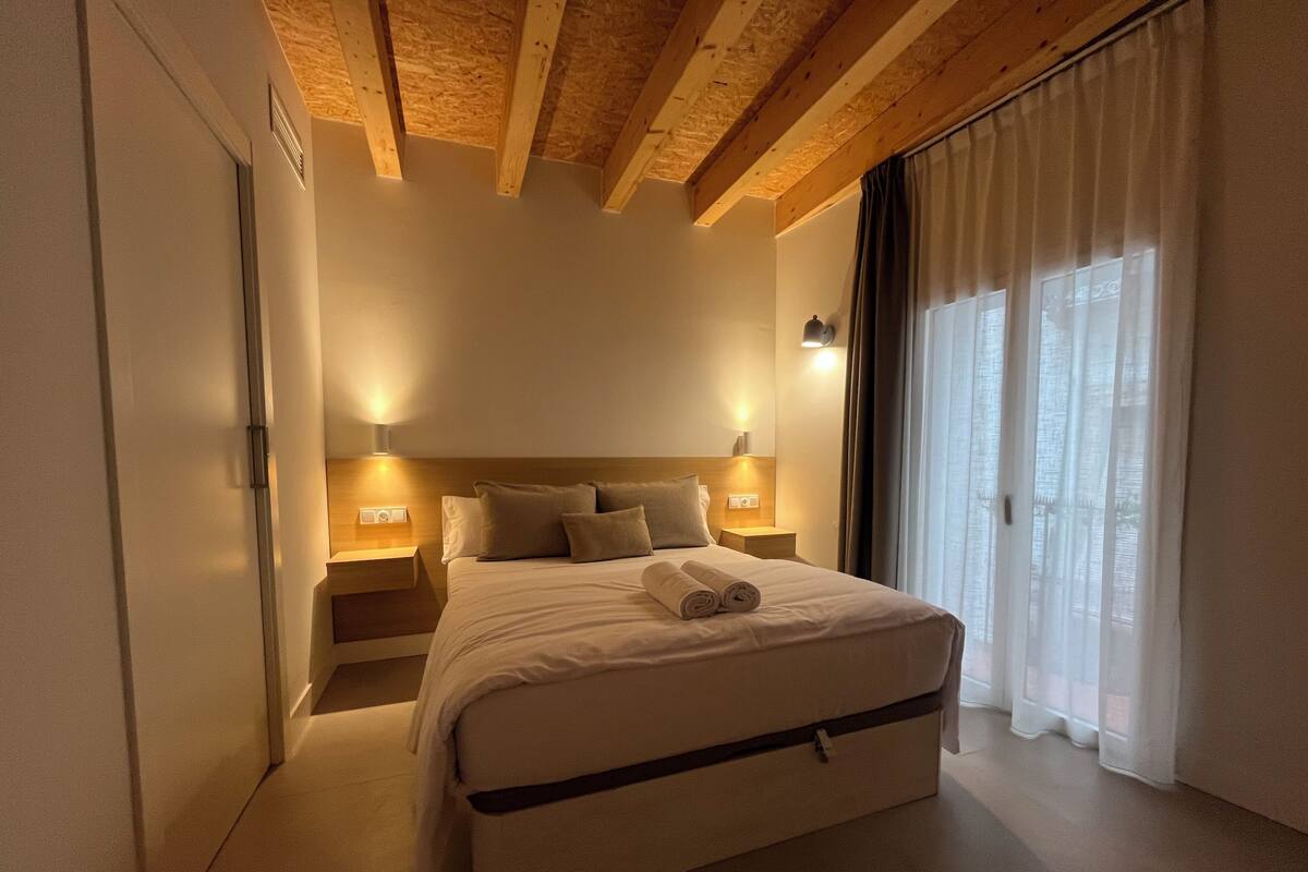 A bedroom with a wooden ceiling, a bed with pillows, a nightstand, and a window with curtains.