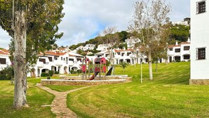 A pathway leading to a playground with a slide and climbing structure, surrounded by white buildings with red roofs and a grassy area. - Holiday Apartment 'Giuliana' with Shared Pool, Wi-Fi, and Air Conditioning (San Jaime Mediterráneo)