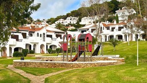 A playground with a slide and climbing structures, surrounded by a stone wall and a paved path. - Holiday Apartment 'Giuliana' with Shared Pool, Wi-Fi, and Air Conditioning (San Jaime Mediterráneo)