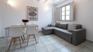 A modern living room with a gray sectional sofa, a dining table with chairs, a vase of red flowers, and a zebra print artwork on the wall. - Oia's Square Residences (Santorini)