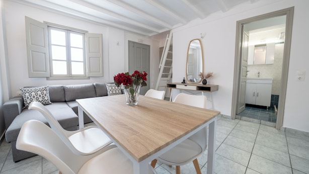 A modern living room with a wooden dining table, white chairs, a gray sofa with patterned cushions, a vase with red flowers, and a large mirror above a white cabinet. - Oia's Square Residences (Santorini)