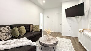 A modern living room with a dark sofa, a round coffee table, and a flat-screen TV mounted on the wall. - Fully Renovated 2BR Executive Residence Near NYC (Cliffside Park)