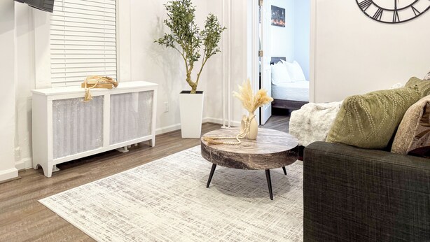 A modern living room with a sofa, a round coffee table, a potted plant, a radiator, and a wall clock. - Fully Renovated 2BR Executive Residence Near NYC (Cliffside Park)