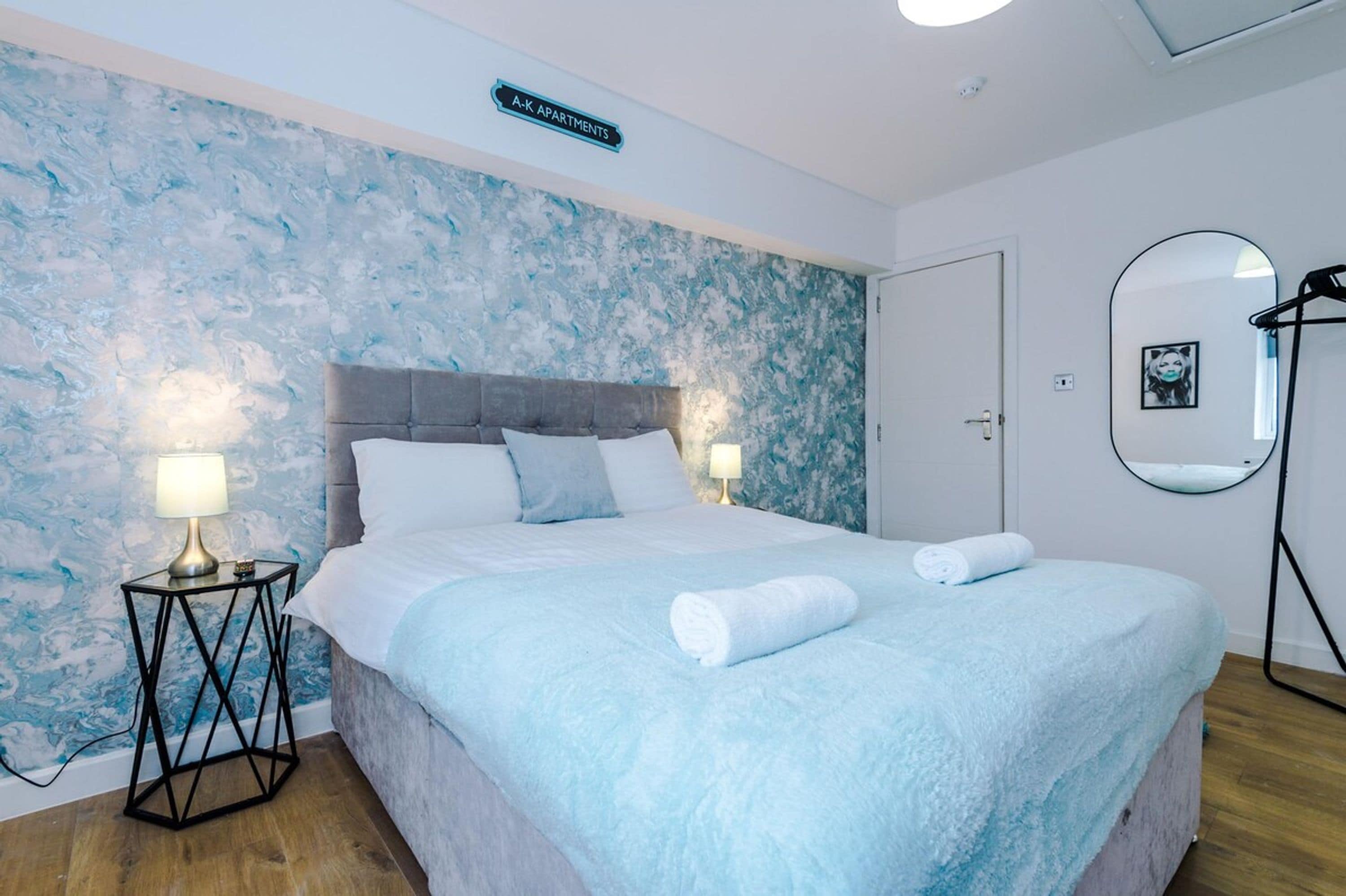 A bedroom with a bed, two bedside lamps, a wall-mounted mirror, and a sign indicating 'A-K Apartments'.